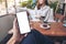 A man`s hand holding black mobile phone with blank white screen with woman sitting in cafe