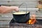 A man`s hand mixes by spoon millet porridge in a pot on the grill with fire, on the wooden fence background