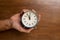 In the man`s hand is an old analog mechanical retro clock. Below is a wooden brown background. The time on the clock is five