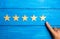 The man`s hand points to the fifth star on a blue wooden background. The concept of assessing quality and status. The critic