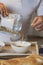 Man's hands emptying granola into a white bowl in a kitchen