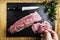 Man`s hands splitting a pork tenderloin with a knife next to some parsley branches on a black slate griddle