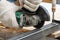 A man in the safety gloves is cutting metal using a green angle grinder tool with sparks on a work bench. Horizontal