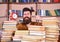 Man, scientist peeking out of piles of books with alarm clock. Teacher or student with beard studying in library. Man on