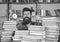 Man, scientist peeking out of piles of books with alarm clock. Teacher or student with beard studying in library. Man on