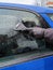 Man scrapes hoarfrost with a plastic scraper from the side window of a blue car. Hand in a black leather glove in the cold.