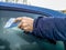 A man scrapes hoarfrost with a plastic scraper from the side window of a blue car. Hand without gloves in the cold. High-rise