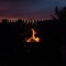 A man in the shade looks at the flames of the fire in the grill.