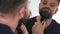 Man shaves his beard using an electric trimmer. Reflection in the mirror