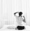 Man in shirt sits on bed, white curtains on background. Guy stretching arms, full of energy in morning, rear view