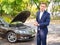 A man shows that he is late standing near a broken car on the road. Outside.