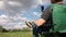 A man sits in a camping chair and photographs the landscape in front of him on the phone. Meadow with green grass. Blue sky with