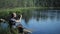 A man sits on a pier of the lake and making photo on the phone. The beautiful blue lake and forest on background