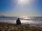 A man sitting by the beach, on a mound of sand, watches the movement of the waves as the sun goes down, in a beautiful blue sky,