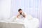 Man sitting on bed, falling asleep while lean on hand, white curtains on background. Handsome tired man leaning on his