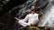 Man sitting in meditation yoga on rock at waterfall in tropical