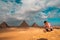 Man sitting on the sandy desert dunes posing in front of the great pyramids of giza. Traveling egypt in winter time, tourists