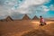 Man sitting on the sandy desert dunes posing in front of the great pyramids of giza. Traveling egypt in winter time, tourists