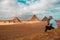 Man sitting on the sandy desert dunes posing in front of the great pyramids of giza. Traveling egypt in winter time, tourists