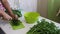 A man slices parsley with a knife on a chopping Board. Puts it in a container. Next to the table there is a lot of parsley and