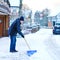 Man with snow shovel cleans sidewalks in winter during snowfall. Winter time in Europe. Young man in warm winter clothes