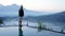 Man standing alone from behind on the edge of the pool at sunrise with water reflection and blue mount Batur view background.