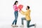 Man standing on knee and gifting bouquet of red paper cut cards with hearts symbols to smiling girl on white background