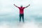 Man standing on rocky outcrop surrounded by white, billowing clouds with his hands reaching the sky