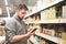 Man stands in the aisle of the supermarket at shelves with canned vegetables and uses a smartphone. Buyer man looks at the