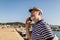 Man stands on pier dressed in a sailor`s shirt and hat using smart phone