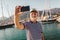 Man stands on pier dressed in a sailor`s shirt and hat using smart phone to make photo