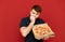 Man stands on a red background with a box of hot pizza and looks at the food and bites his fingers from the nerves. Man with a