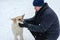 A man is stroking a large street dog in the snow. An adult man crouches next to a white and red male dog. A kind stray dog. A