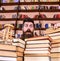 Man on surprised face between piles of books, while studying in library, bookshelves on background. Teacher or student