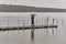 Man talking on phone and walking under umbrella on wooden pier on rainy gray day over of backdrop of lake and forest.