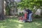 Man with tattoos on standing commercial lawnmower cutting a close circle around tree and flowerbeds in upscale