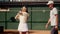 A man tennis coach comes closer her ward and shows how to hold racket properly on shoulder to repel a blow shot of rival