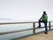 Man thinking. Tourist in green on sea mole at handrail. Autumn mist