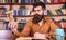 Man on thoughtful face holds hourglass while studying, bookshelves on background. Teacher or student with beard studying