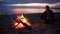 Man is throwing firewood at a fire on the river bank at sunset