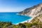 Man tourist standing on top of a rock, raising hands with an exciting feeling of freedom, looking at Myrtos Beach