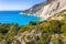 Man tourist standing on top of a rock, raising hands with an exciting feeling of freedom, looking at Myrtos Beach