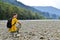 Man tourist in yellow t-shirt with a backpack sitting on the rocks on the dried river, in background mountains and river