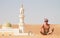 Man traveling in Oman, topless wearing a turban and shorts, sitting on send dune in fron of mosque in desert Oman
