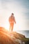 Man traveller with backpack stands on top of rock cliff in grass and looking at sea landscape at sunset, freedom of adventure