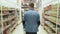 Man with trolley goes among shelves with goods in supermarket, backside view, steadicam shot.