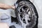 A man use a large brush to scrub clean a car\'s front tire and loosen dirt and grime in the grooves.