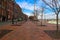 A man walking down a red brick sidewalk along the riverfront with rows of bare trees and tall red brick buildings and parked cars