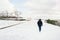 A man walks along an empty snow covered promenade during the day. The clouds are white. There are trees on one side and the sea on