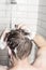 A man washes his hair with shampoo in the bathroom, against the background of a shower with running water. Back view